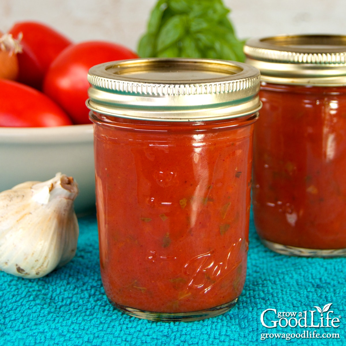 Jars of home canned pizza sauce on a blue towel surrounded by garlic, ripe tomatoes, and fresh basil.