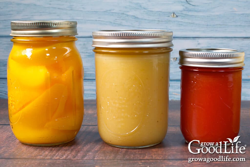 Jars of home canned peaches, applesauce, and strawberry jelly on a table.