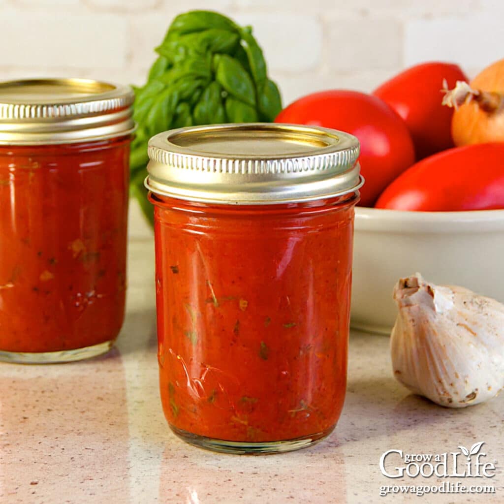 Jars of home canned roasted tomato sauce on a kitchen counter surrounded by garlic, ripe tomatoes, and fresh basil.