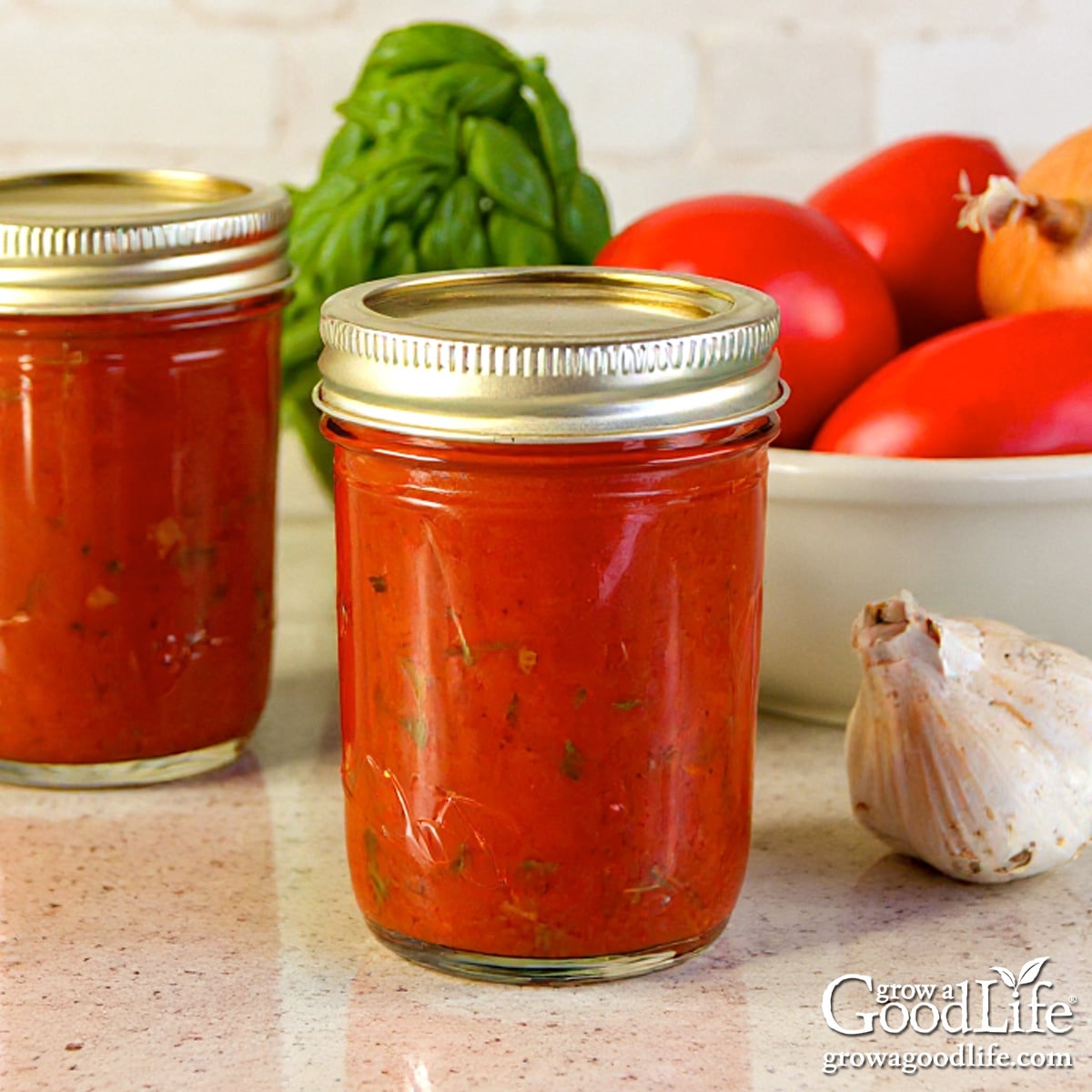 Jars of home canned roasted tomato sauce on a kitchen counter surrounded by garlic, ripe tomatoes, and fresh basil.