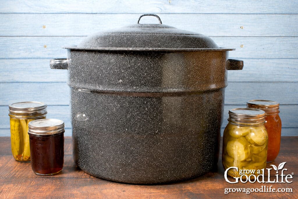Boiling water canner on a table surrounded by jars of pickles, preserves, and other water bath canned foods.