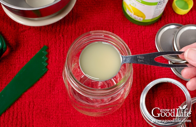 Hand holding a tablespoon of bottled lemon juice over an empty canning jar before adding tomato sauce.